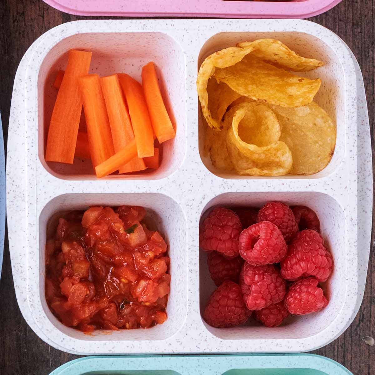 A four compartment snack box containing carrot battens, potato chips, salsa and raspberries.