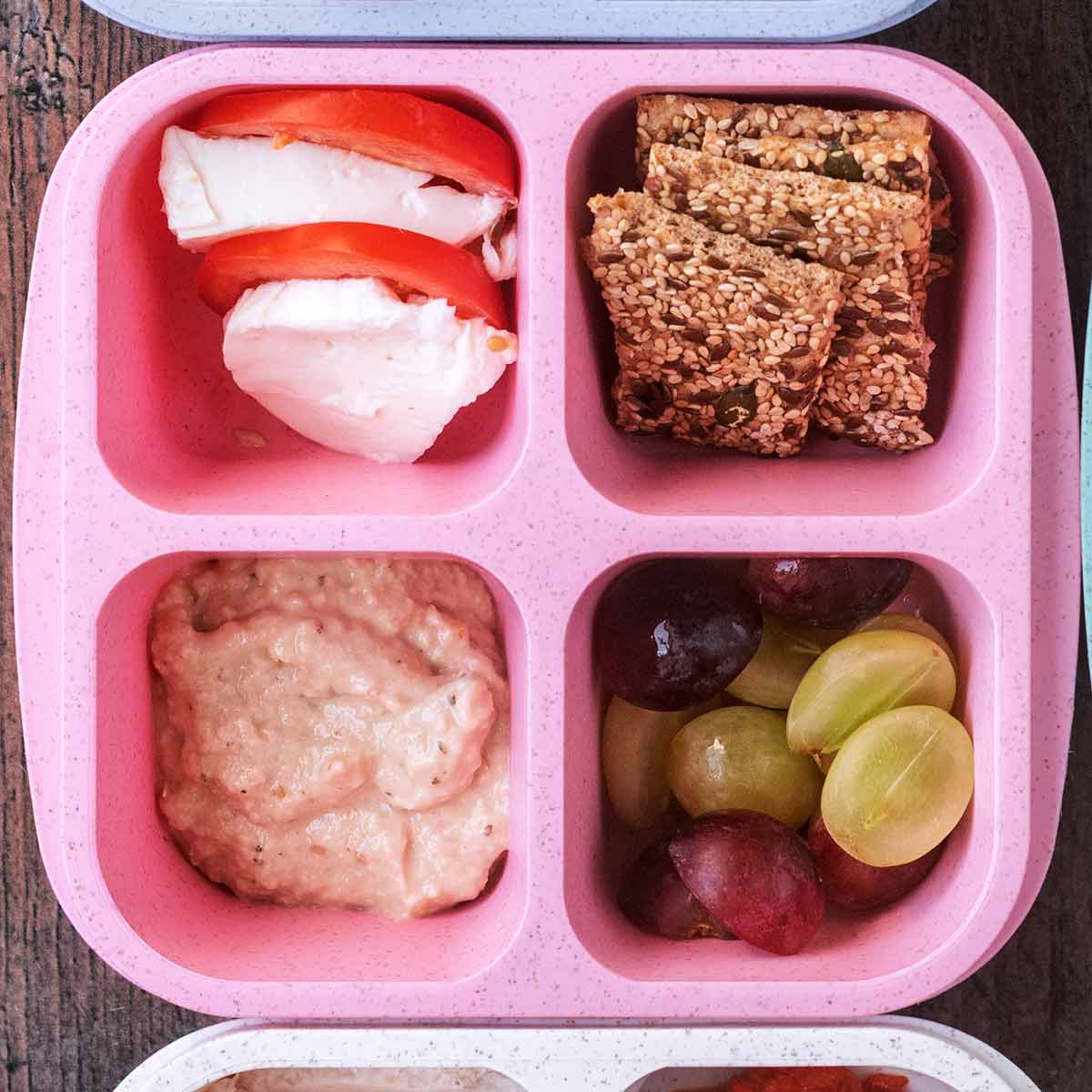 A four compartment snack box containing tomato slices with mozzarella, seeded crackers, baba ganoush and grapes.