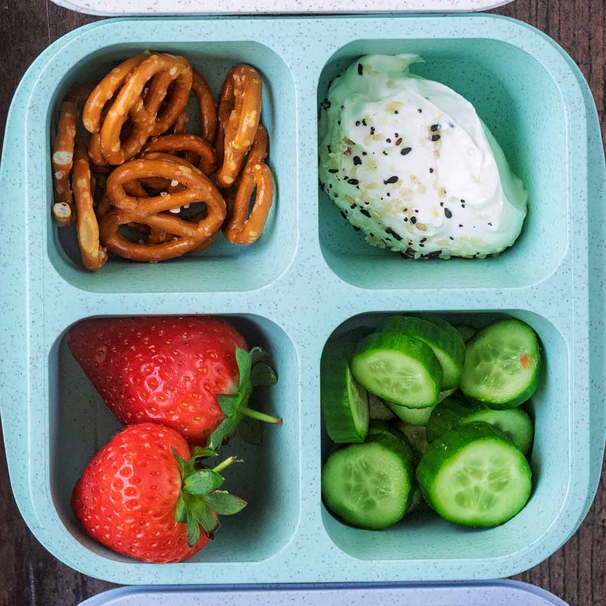 A four compartment snack box containing mini pretzels, cream cheese, cucumber slices and strawberries.