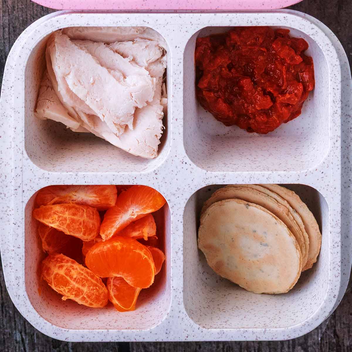 A four compartment snack box containing cooked chicken slices, pepper dip, crackers and orange segments.