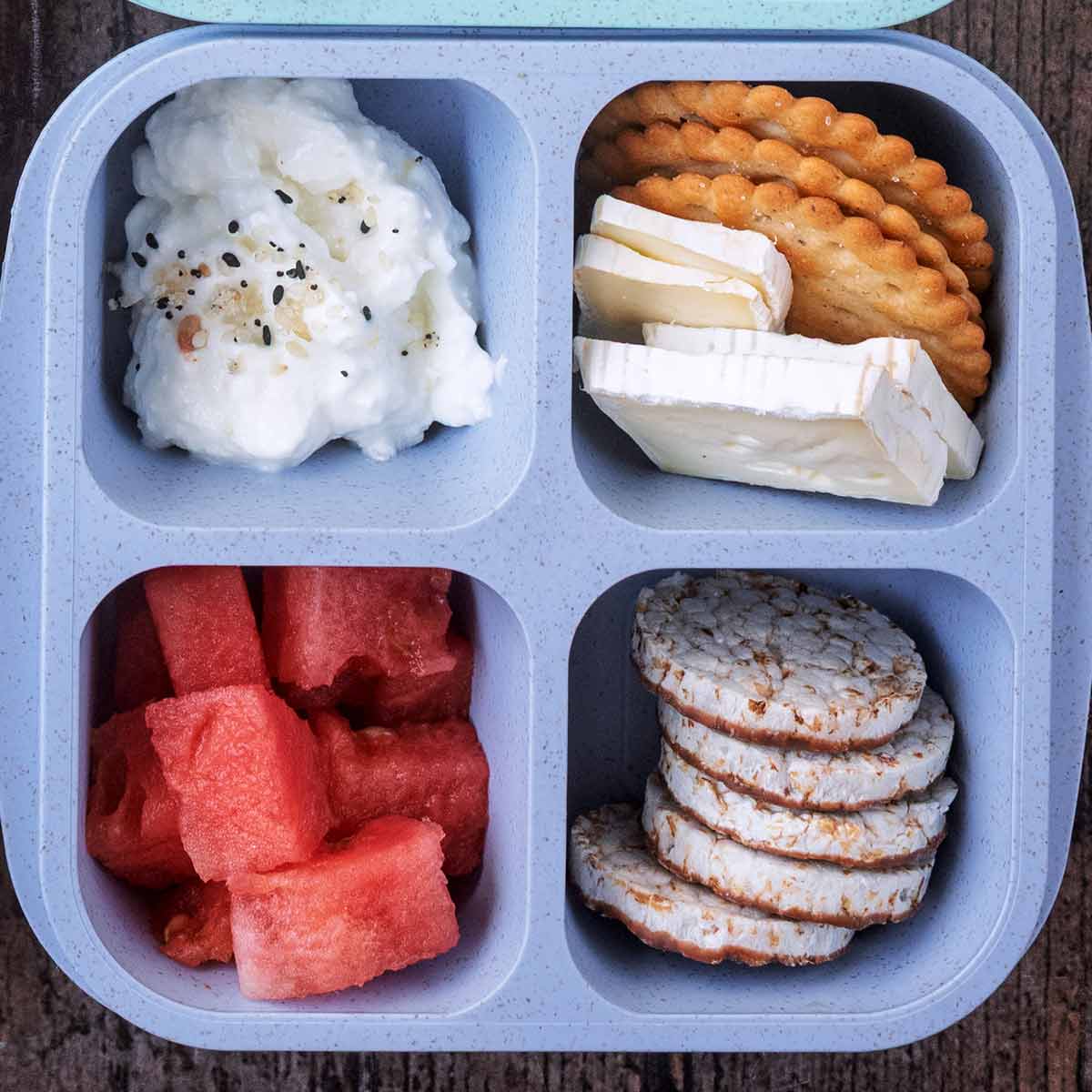 A four compartment snack box containing cottage cheese, brie with crackers, mini rice cakes and cubes of watermelon.