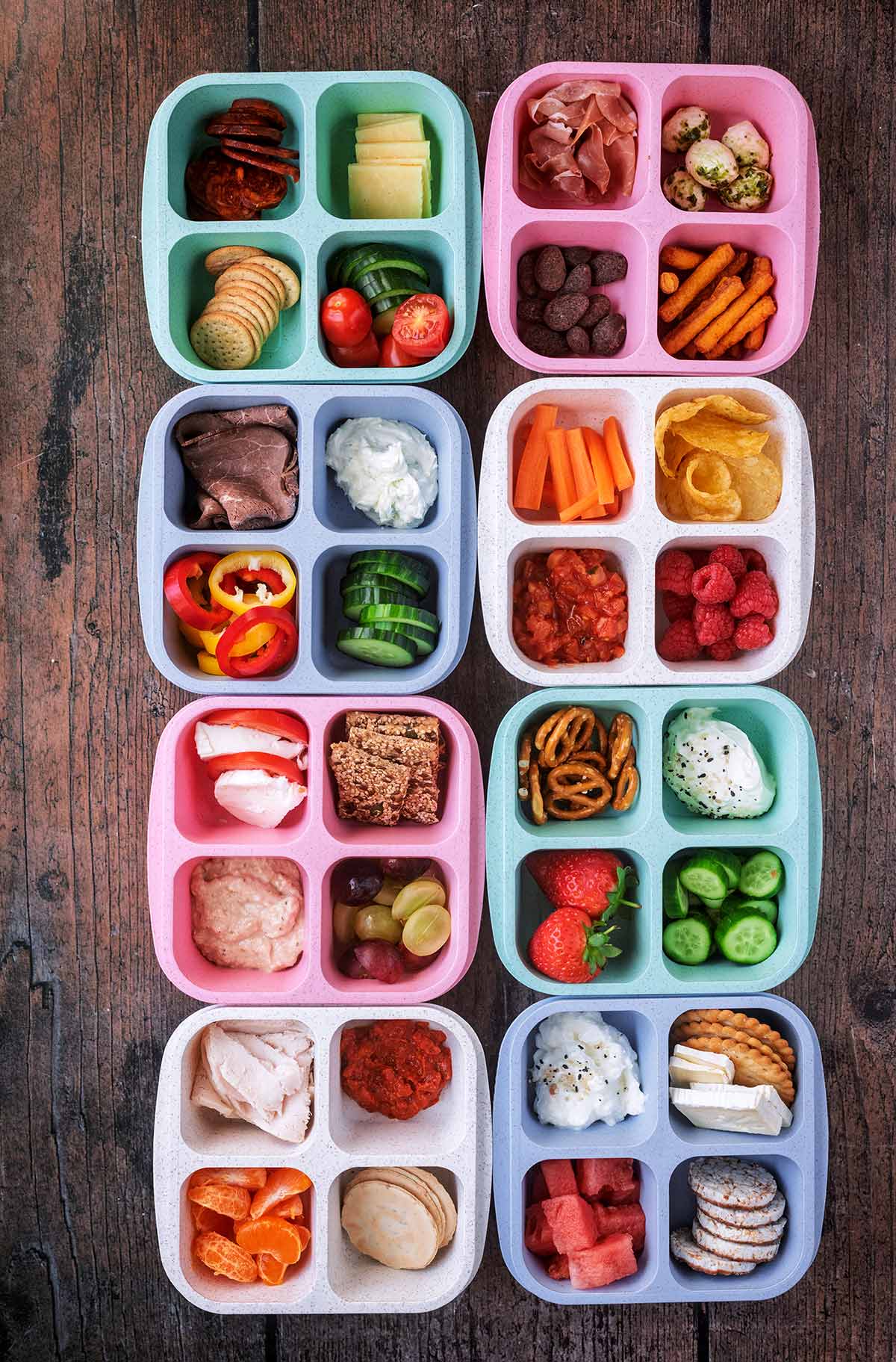 Eight four-compartment snack boxes each containing different meats, vegetables, fruits, crackers and dips.