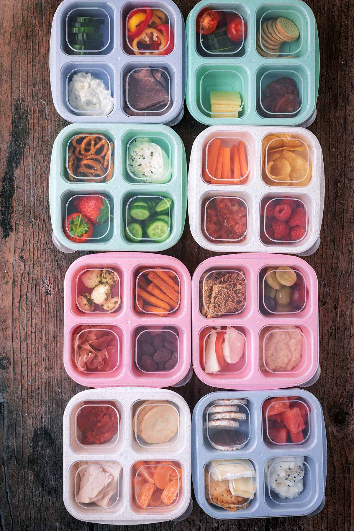 Eight four-compartment snack boxes with clear lids, each containing different foods.