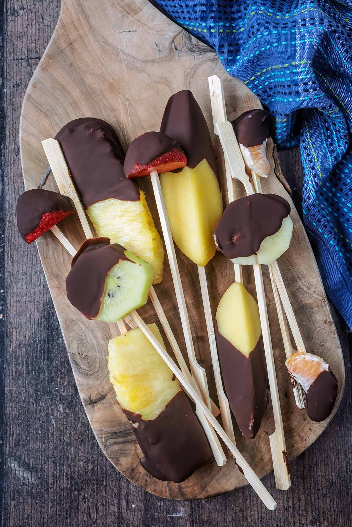 Chocolate covered pieces of fruit on skewers on a wooden board.
