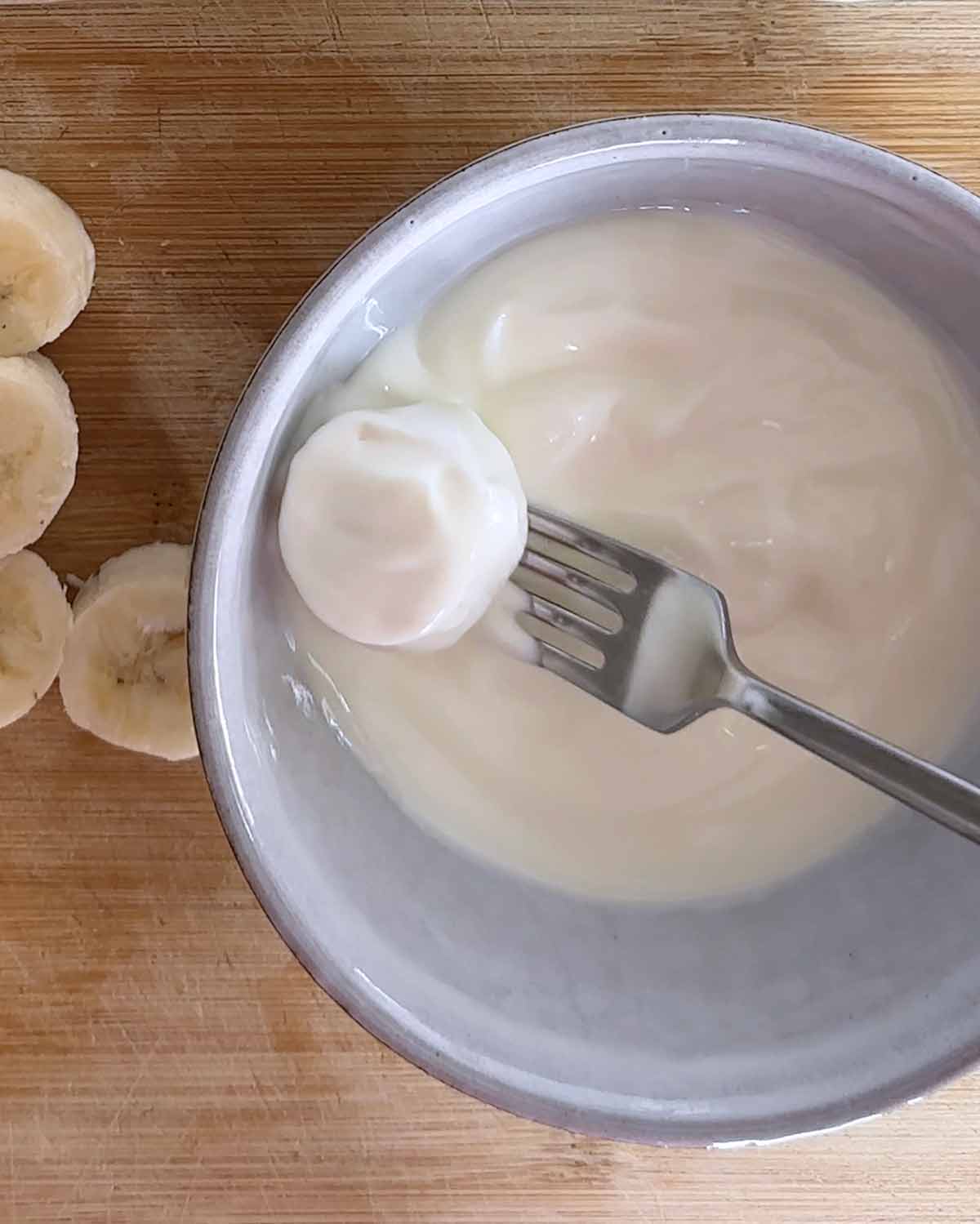 A fork holding a banana slice in a bowl of yogurt.