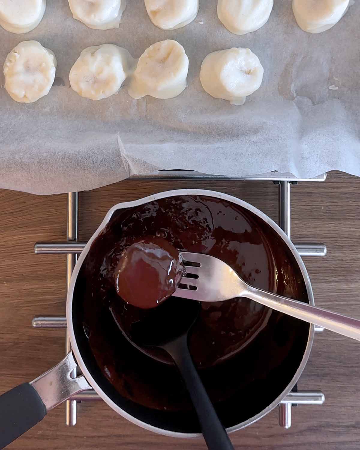 A fork dipping the frozen yogurt covered banana slice into melted chocolate.