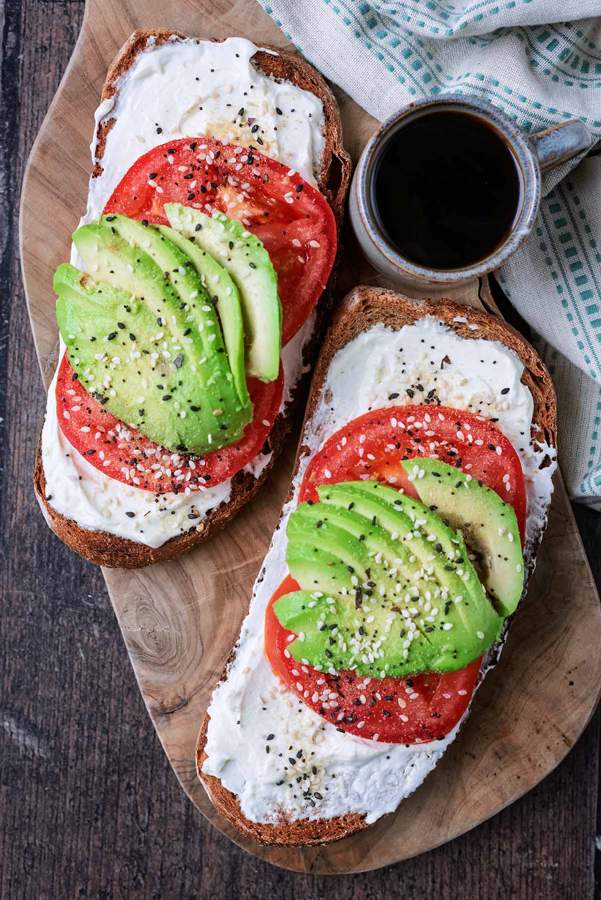 Two sliced of toast topped with cream cheese, tomato slices, sliced avocado and seasoning.