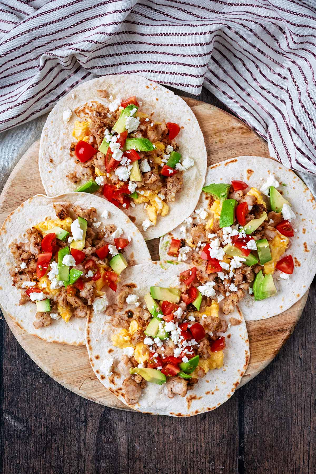 Four sausage, egg and vegetable tacos on a wooden serving board.