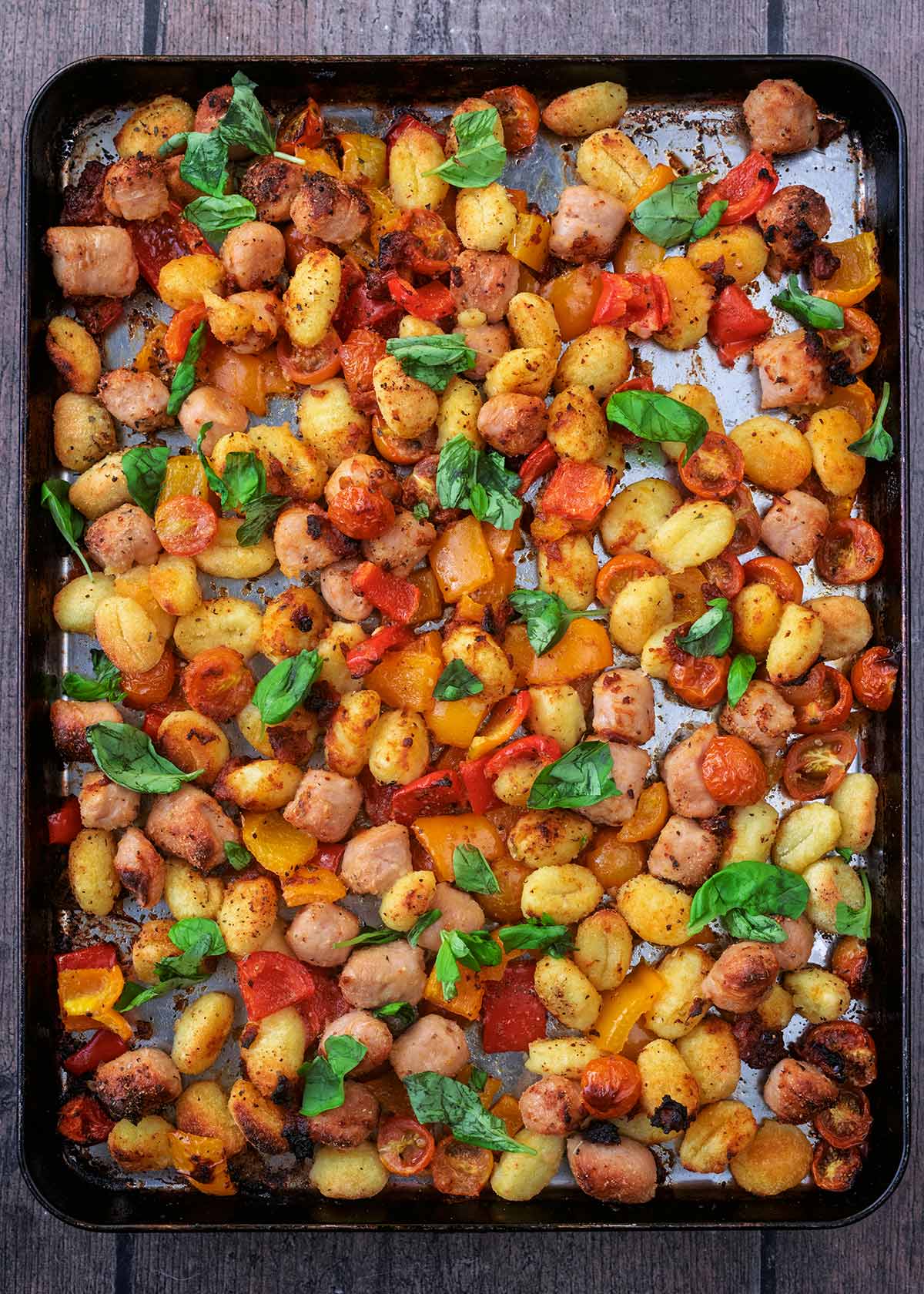 A large baking tray full of cooked chicken sausages, vegetables and gnocchi.