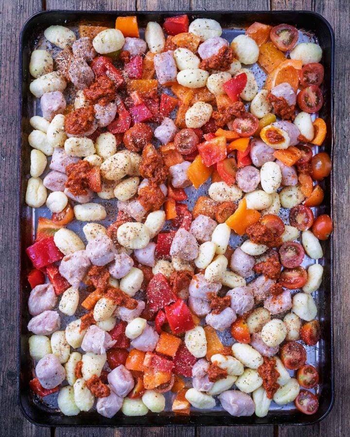 Uncooked gnocchi, diced bell peppers, chunks of sausage meat and dots of pesto on a large baking tray.