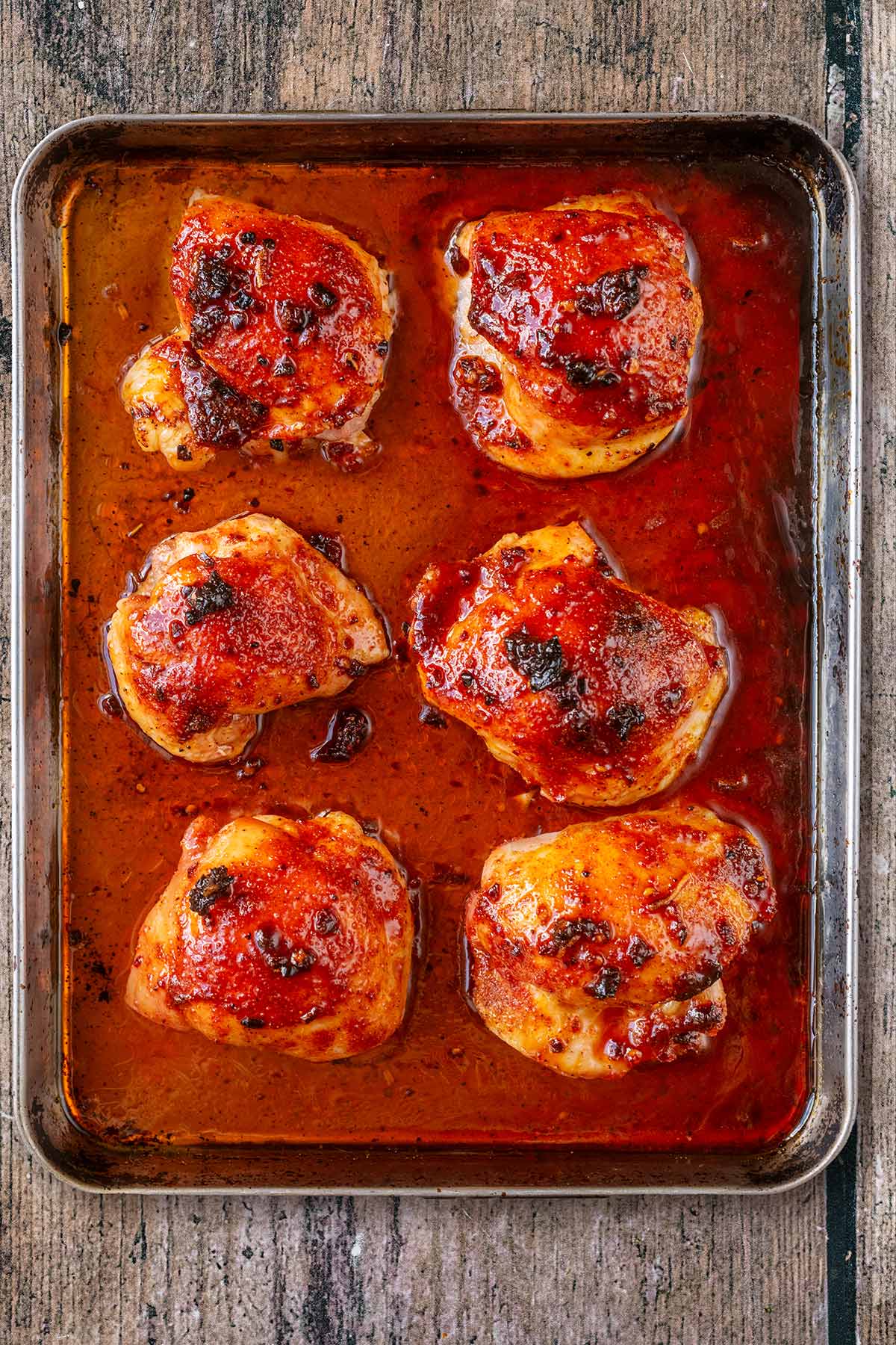 Six baked chicken thighs in a red coloured sauce on a baking tray.