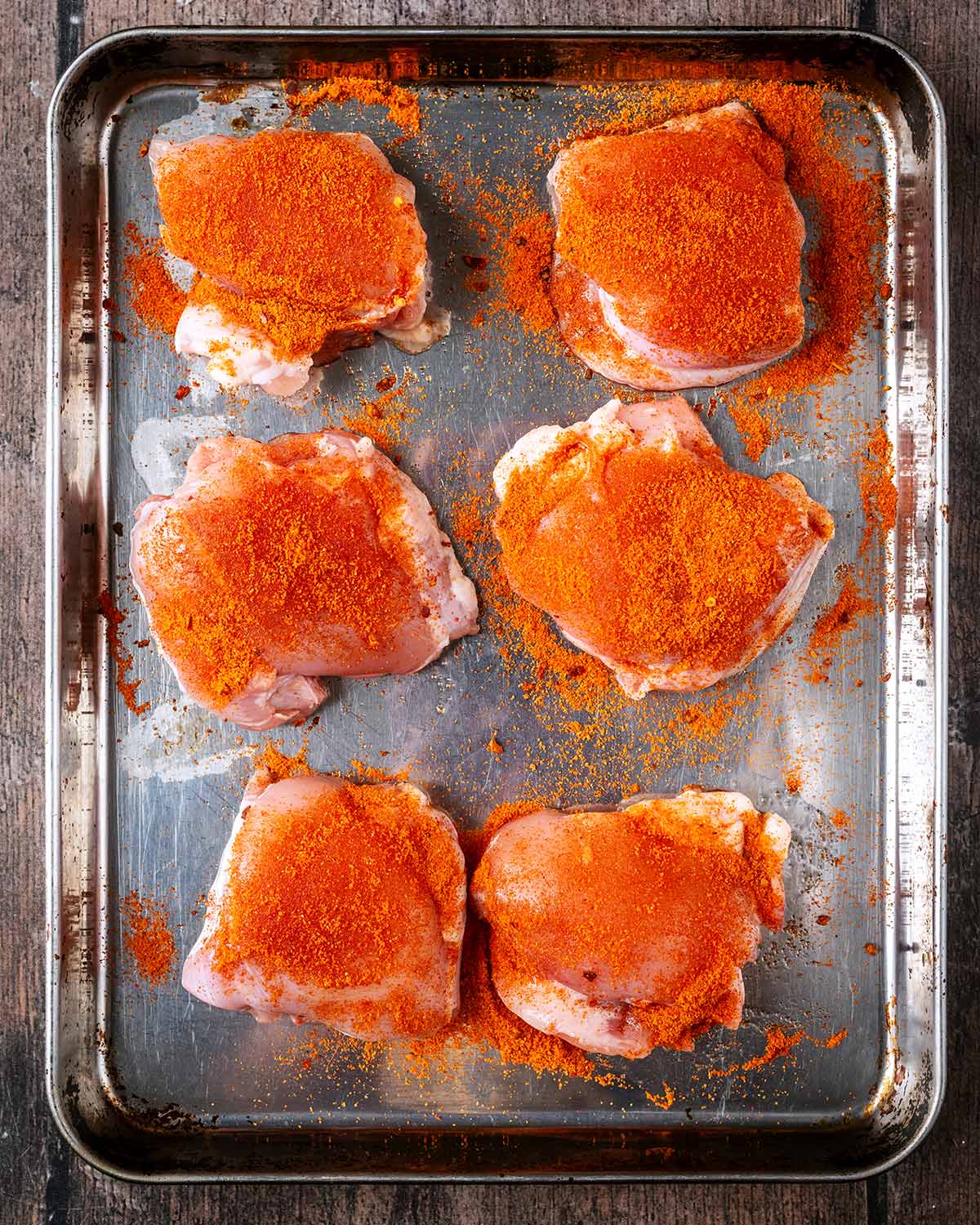 Six raw chicken thighs coated in a seasoning on a baking tray.