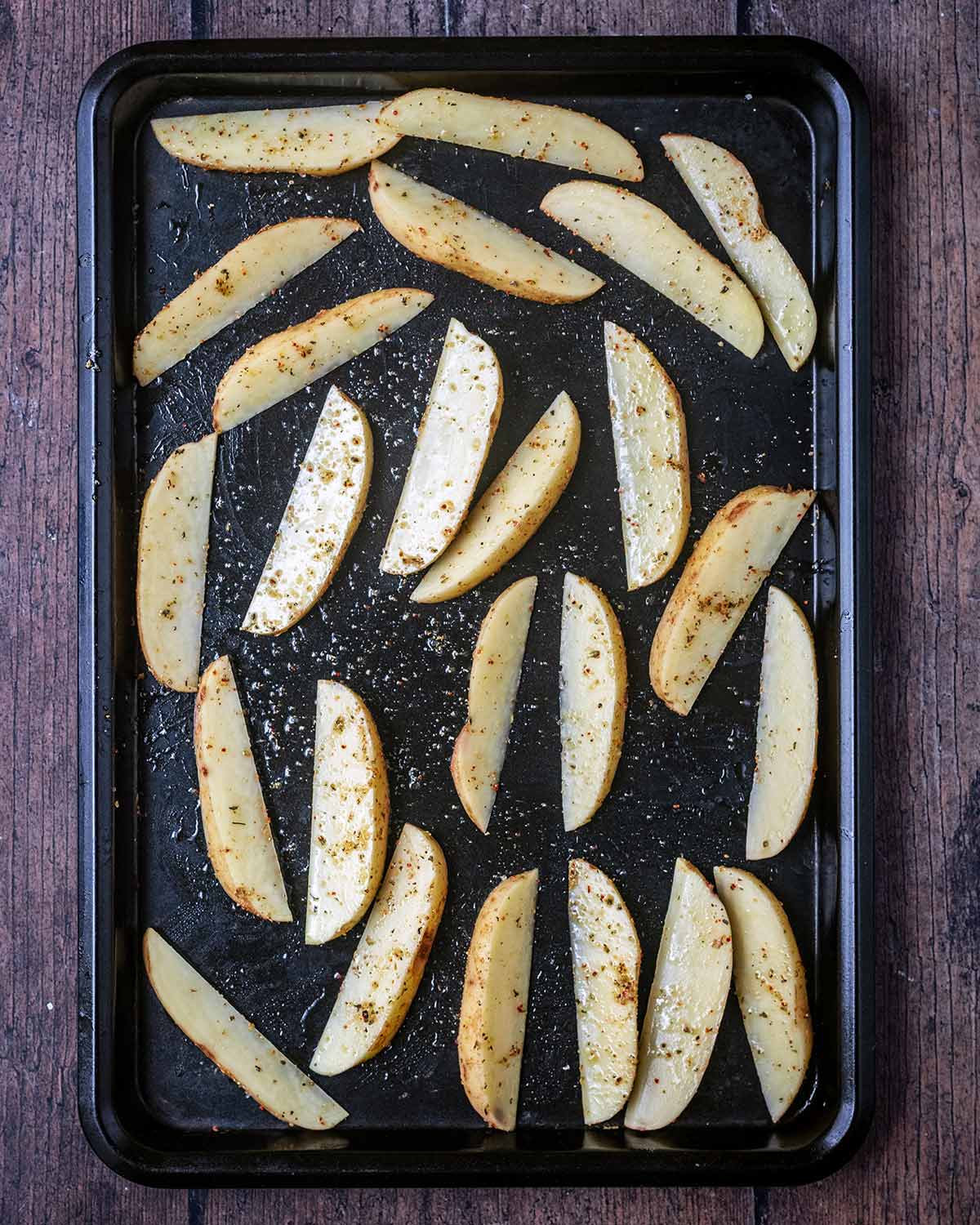 Seasoned potato wedges spread over a large black baking tray.