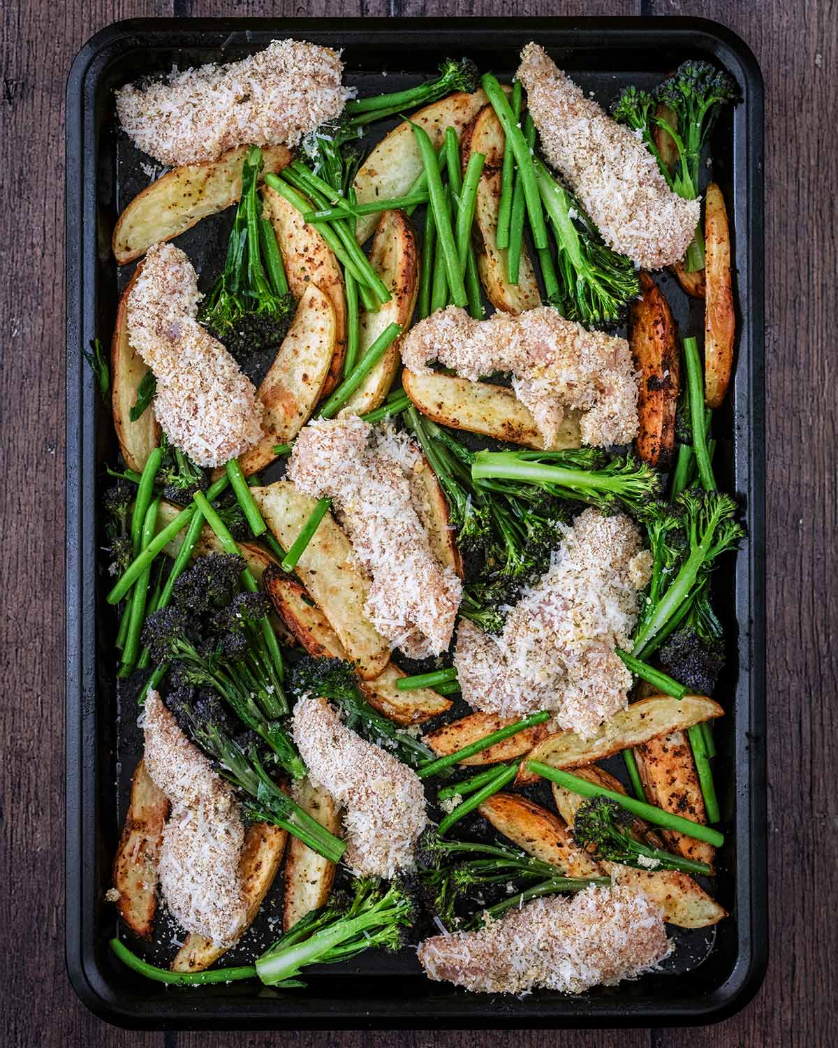 Breaded chicken, part cooked potato wedges, broccoli and beans on a baking tray.