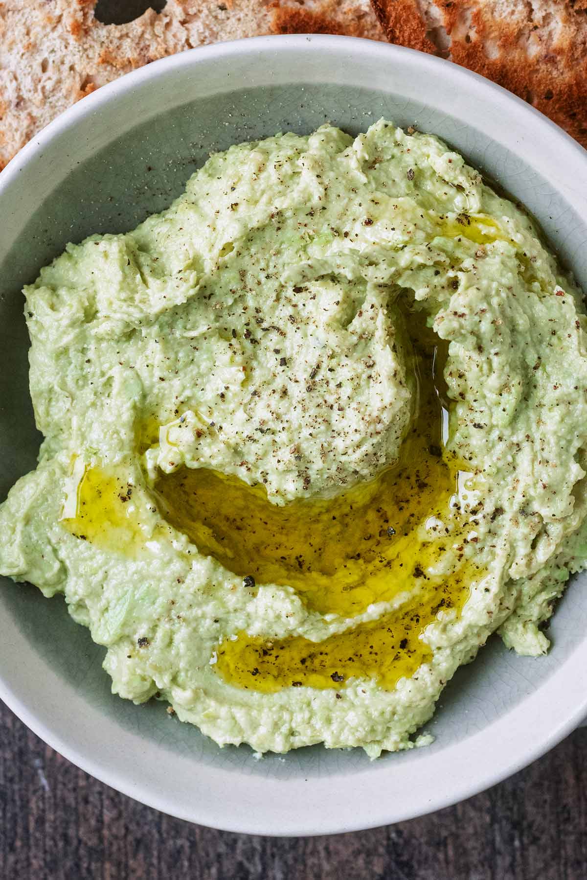 A green coloured hummus in a round bowl with olive oil drizzled on top.