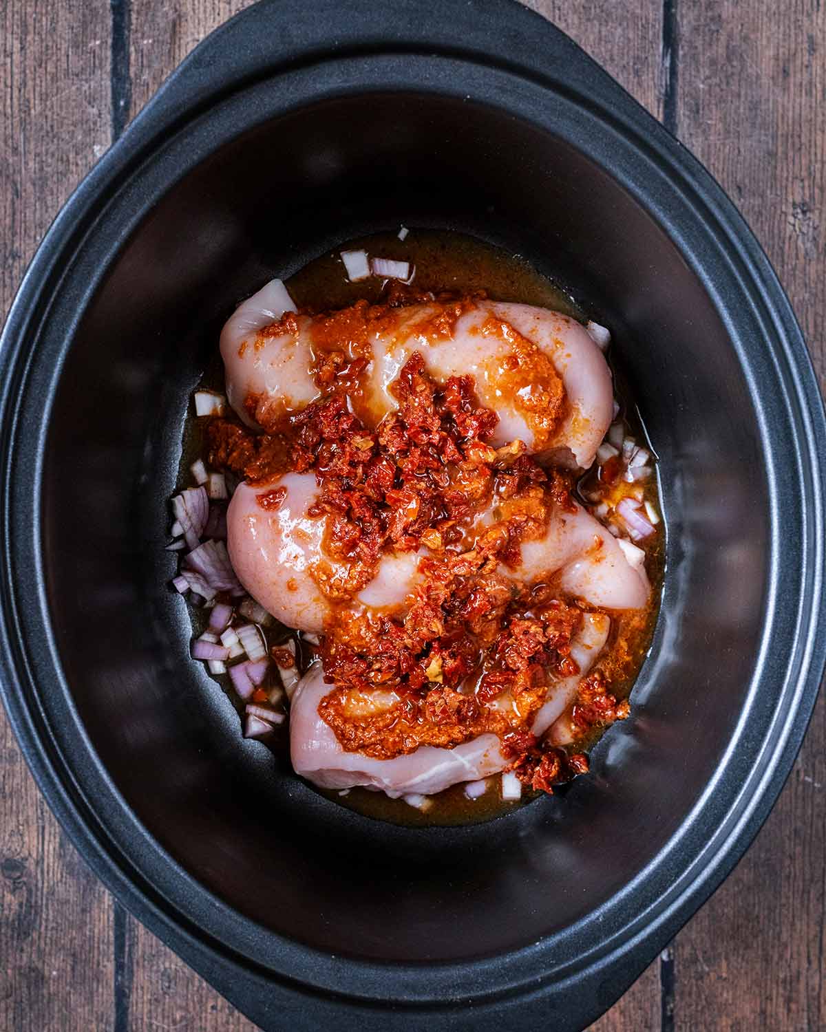 Raw chicken breasts, chopped onion, sun-dried tomatoes and red pesto in a slow cooker pot.