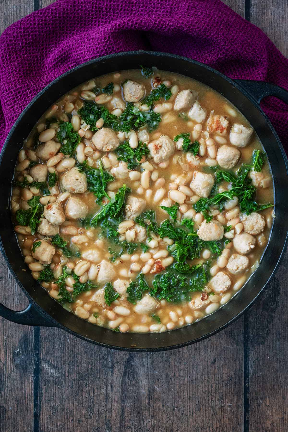 A large black pan with white beans, chunks of sausage and kale in it.