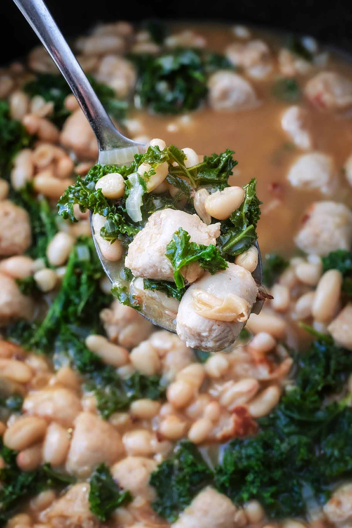 A spoon lifting slices of sausage, beans and kale from a pan.