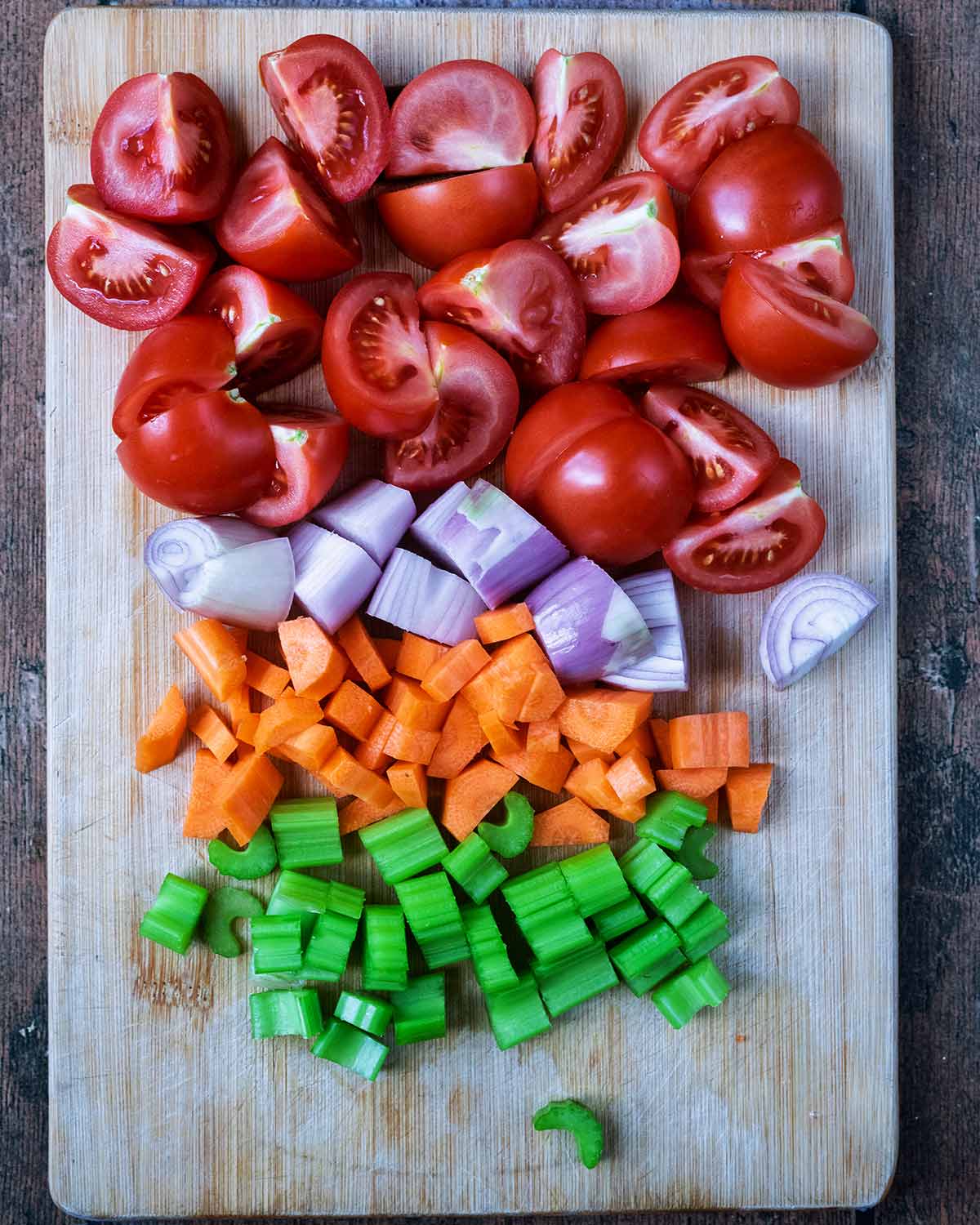A wooden chopping board with tomatoes, celery, carrots and shallots on it.