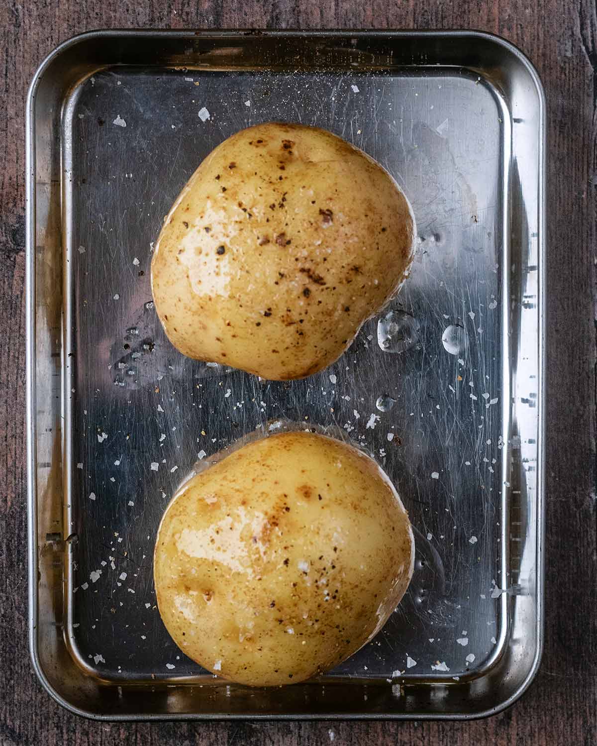 Two large potatoes covered in oil, salt and pepper on a baking tray.