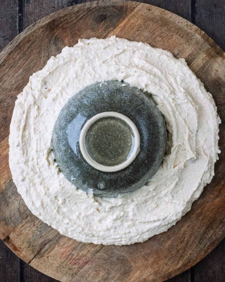 An upturned bowl surrounded by hummus on a wooden board.
