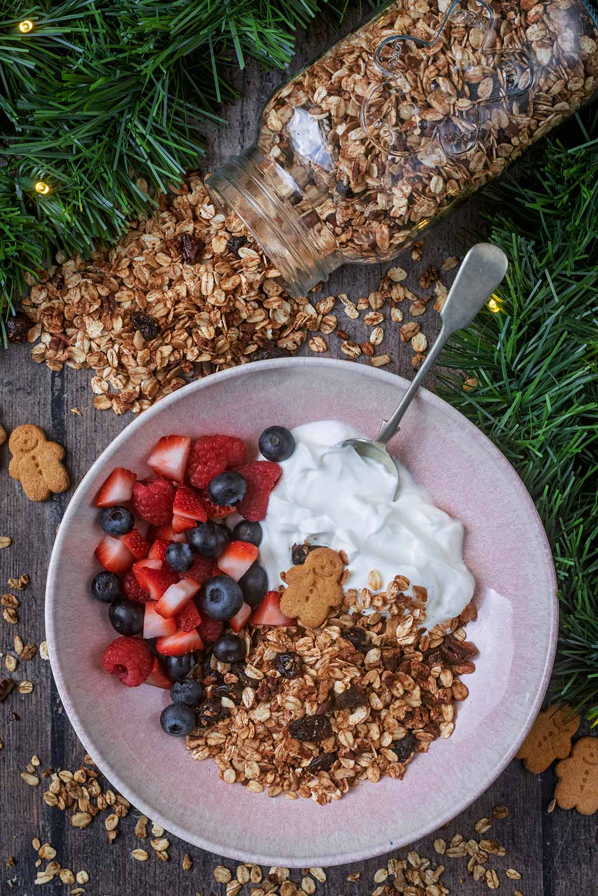 A bowl of granola, yogurt and berries next to a jar that has spilled granola over the wooden surface.