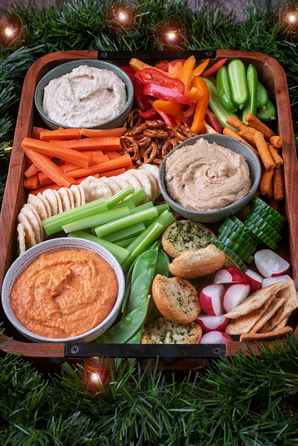 A wooden tray with bowls of hummus, vegetables and crackers.