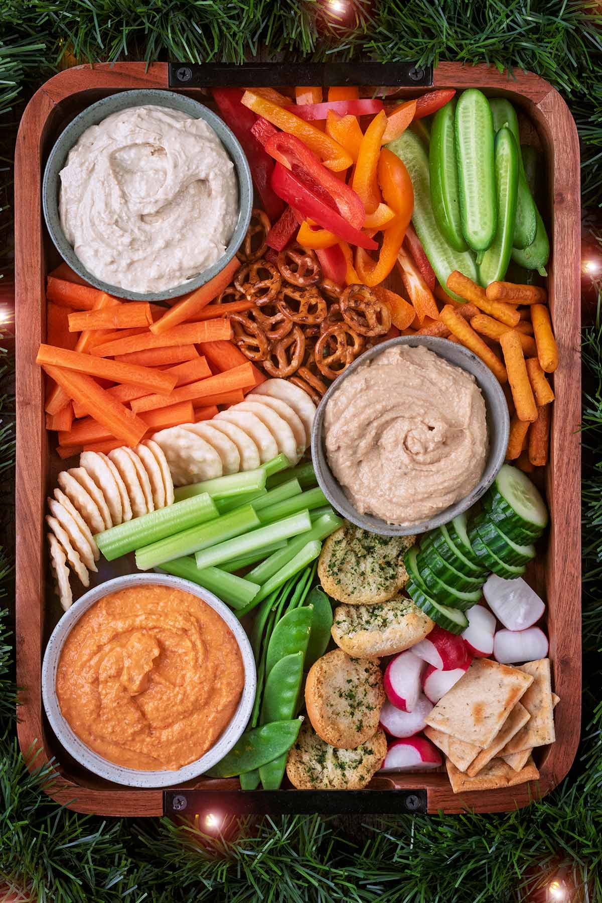 A wooden serving board filled with three types of hummus, crudit&eacute;s and breads.