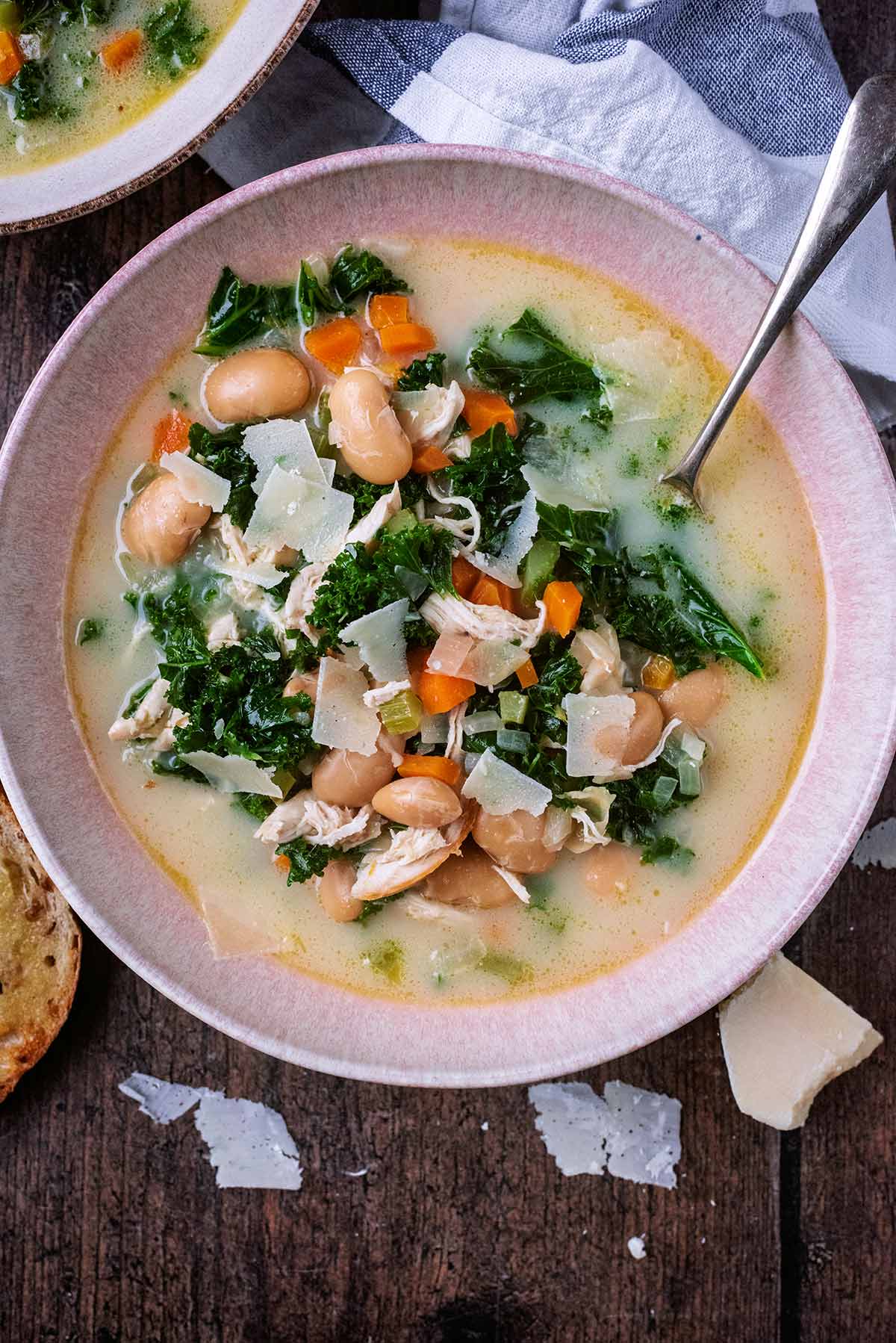A bowl of chicken and bean soup with Parmesan shavings next to it.