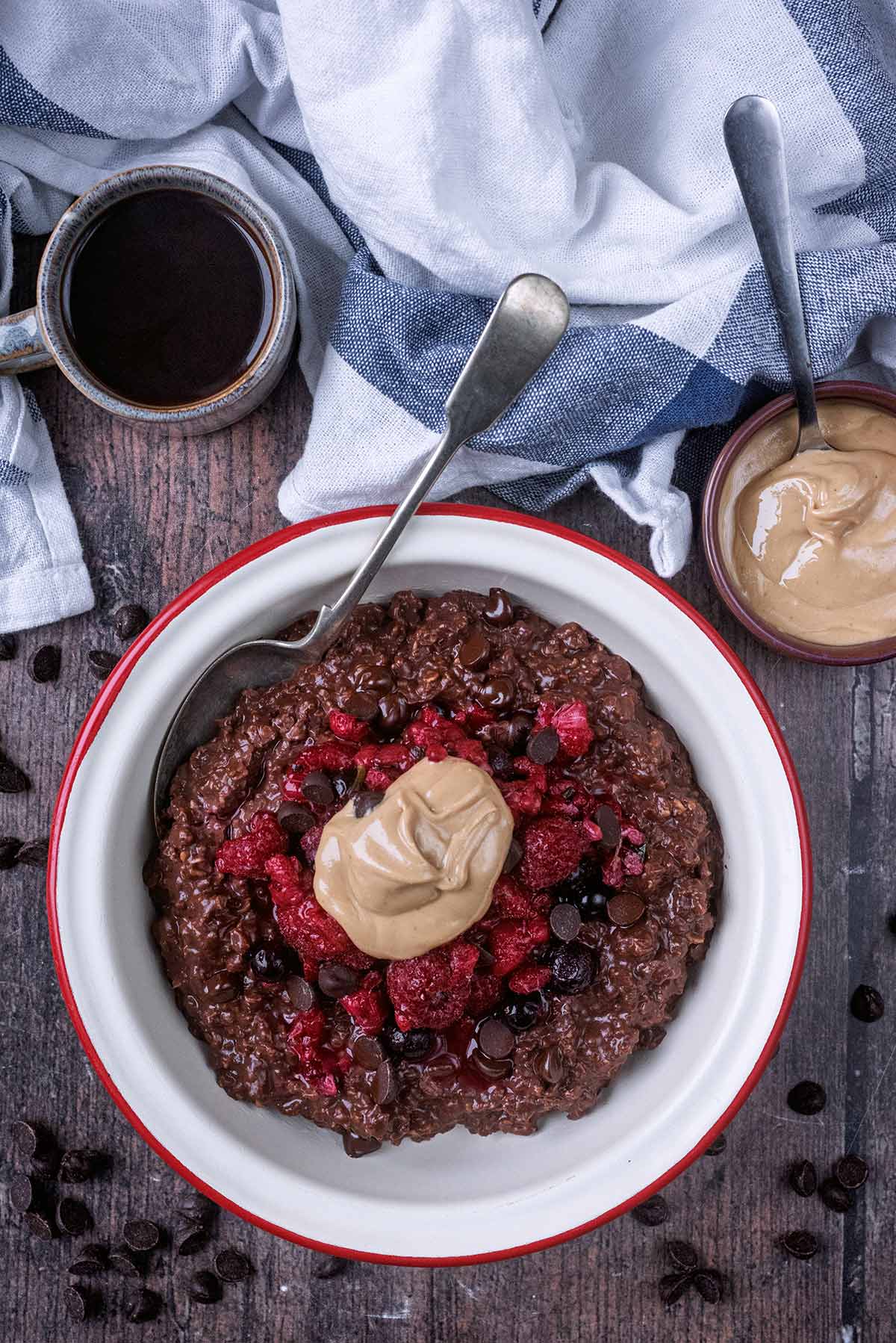 A bowl of chocolate porridge next to a small bowl of nut butter and a cup of coffee.