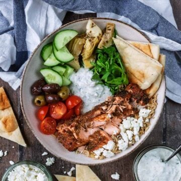 A greek salmon bowl next to a striped towel, cut flatbread, feta and tzatziki.