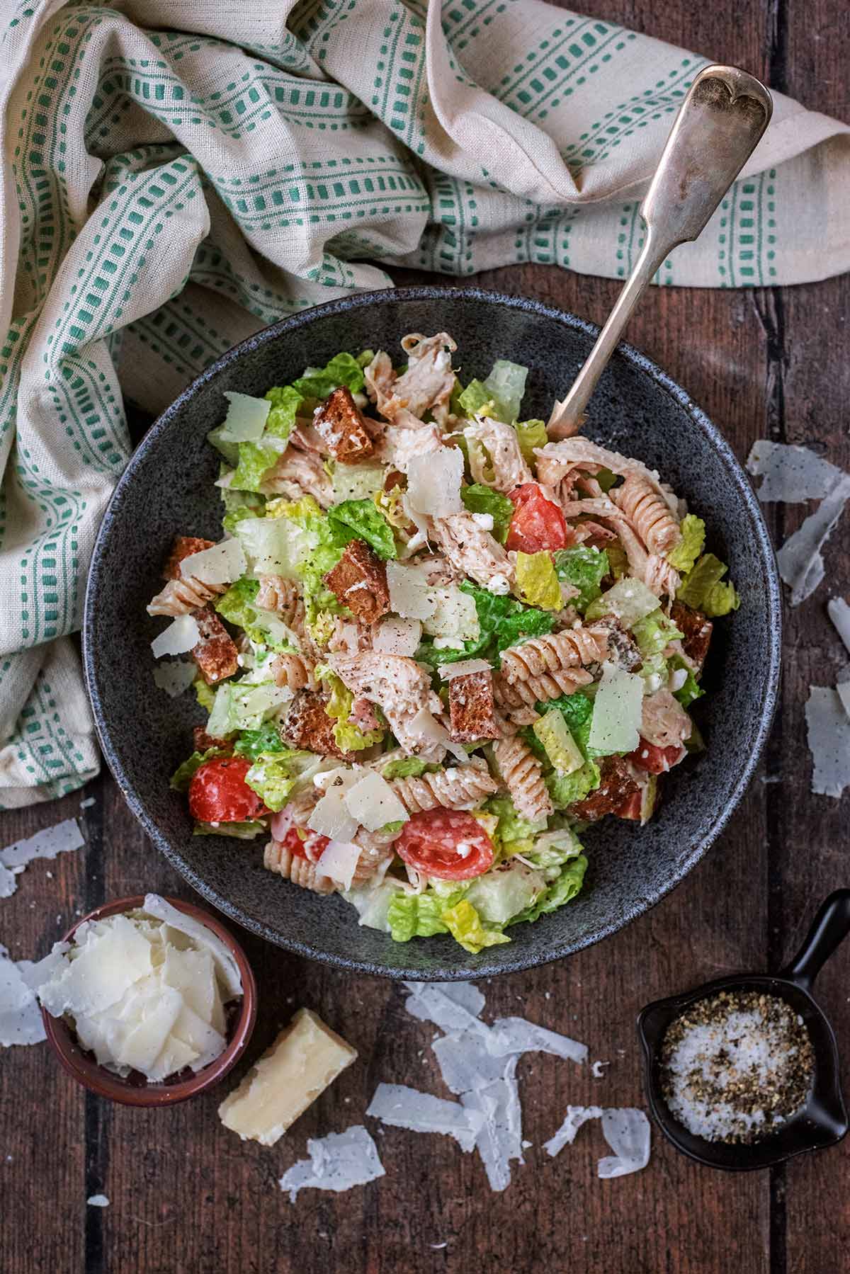 A bowl of pasta salad next to a patterned towel and Parmesan shavings.