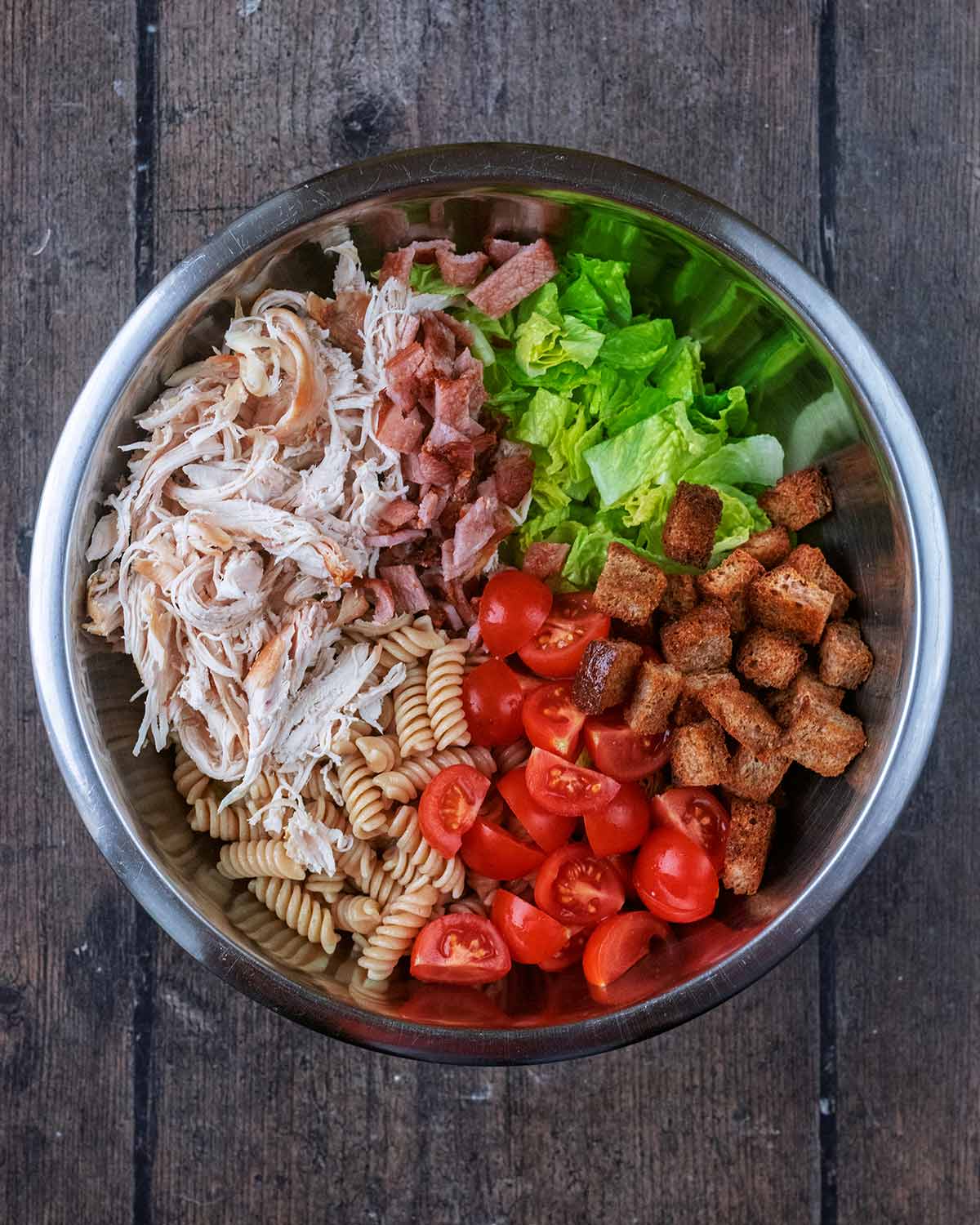 A large bowl with chopped lettuce, tomatoes, pasta, shredded chicken and chopped bacon.