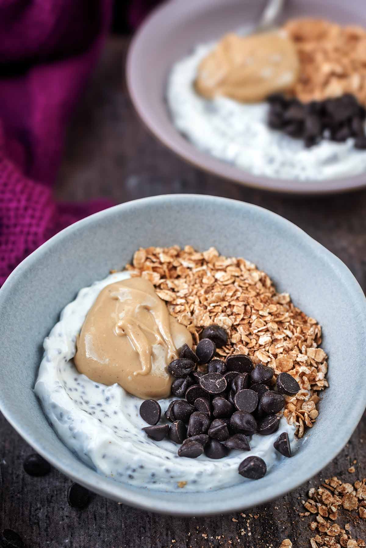 A bowl of yogurt, oats, nut butter and chocolate chips, with another bowl of the same in the background.
