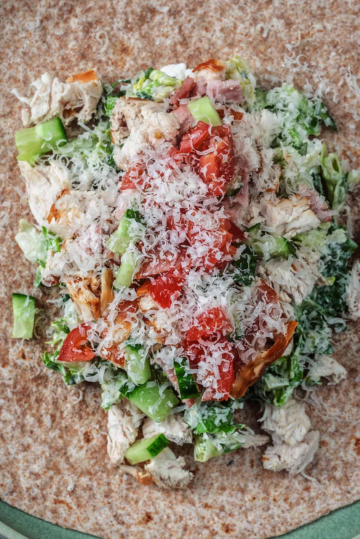 A wholewheat wrap topped with chopped dressed salad, chicken, bacon, salad veg and grated cheese.