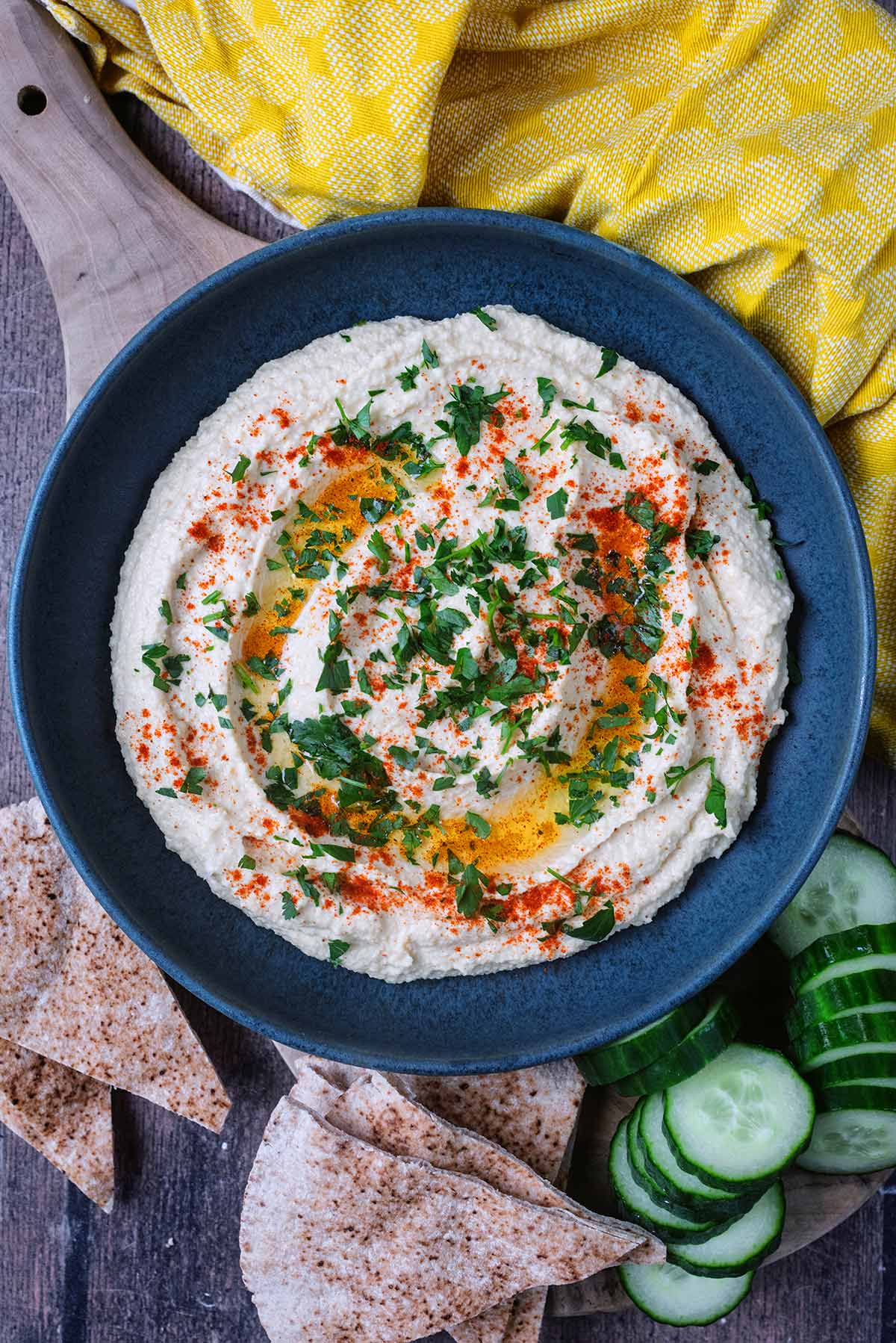 A large bowl full of hummus next to pita bread and slices of cucumber.