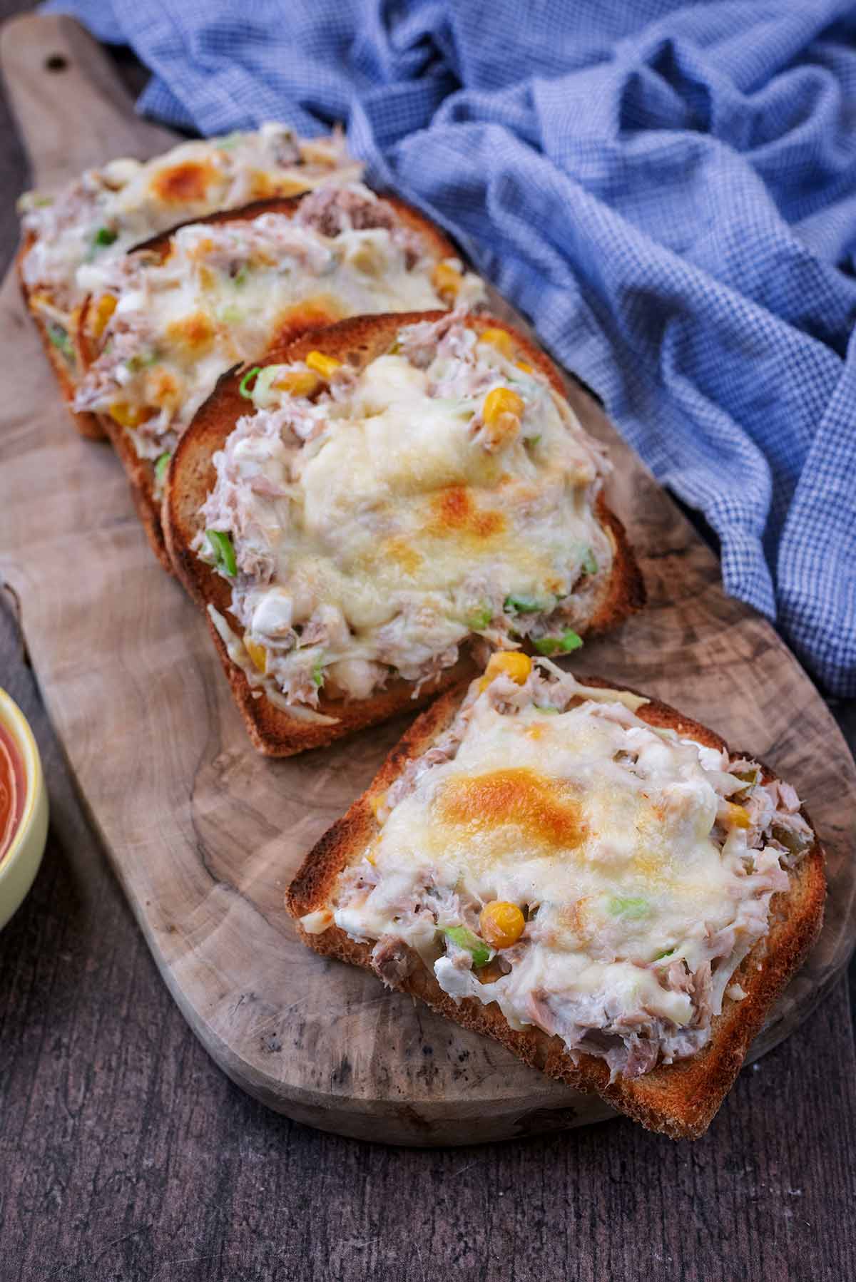 Four tuna melts on a wooden board in front of a blue towel.