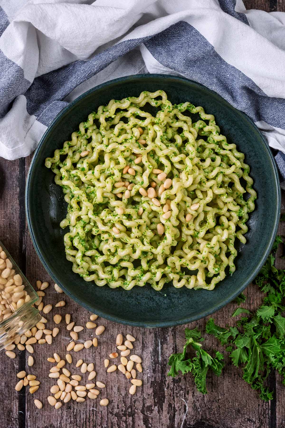 A bowl of pesto pasta next to a jar of knocked over pine nuts.