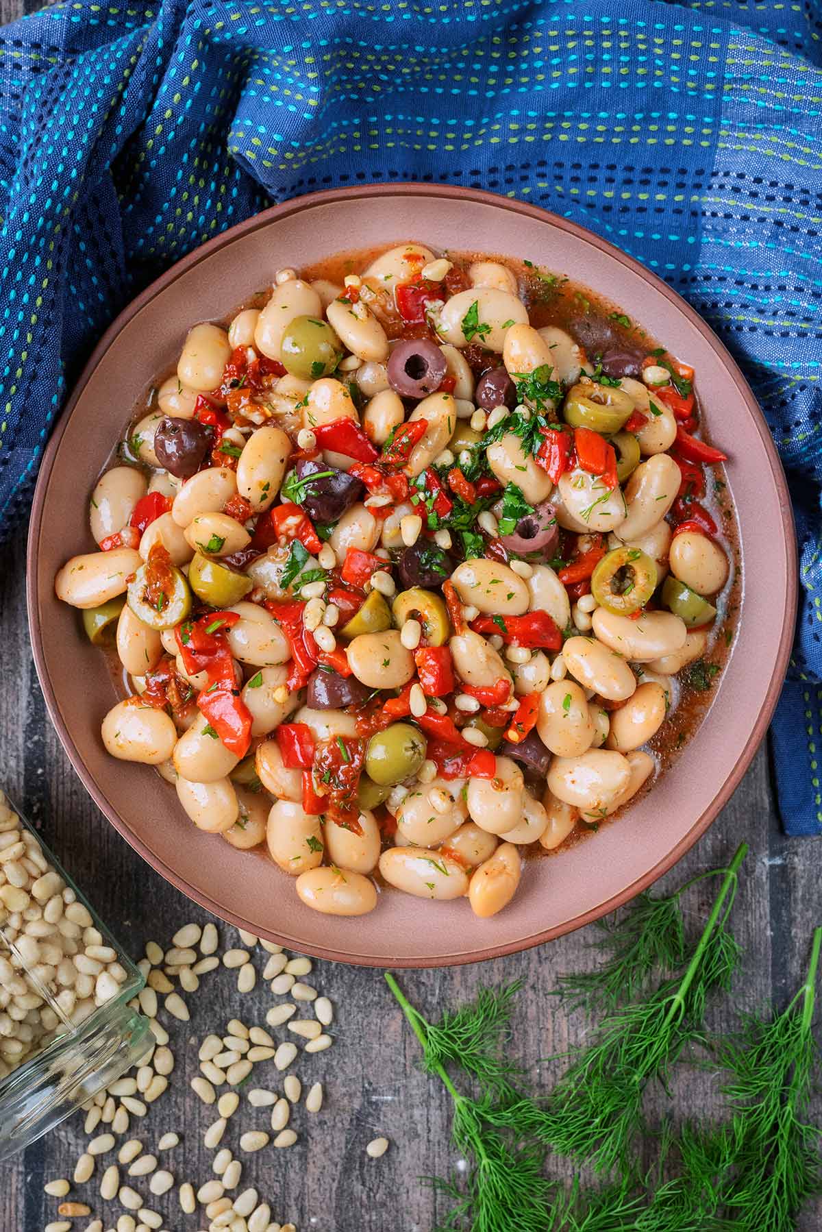 A bowl containing butter bean salad next to a blue towel, fresh dill and pine nuts.