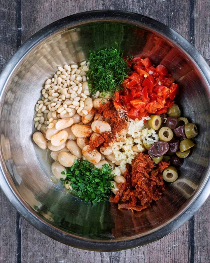 A large mixing bowl containing beans, olives, herbs, tomatoes, garlic and pine nuts.
