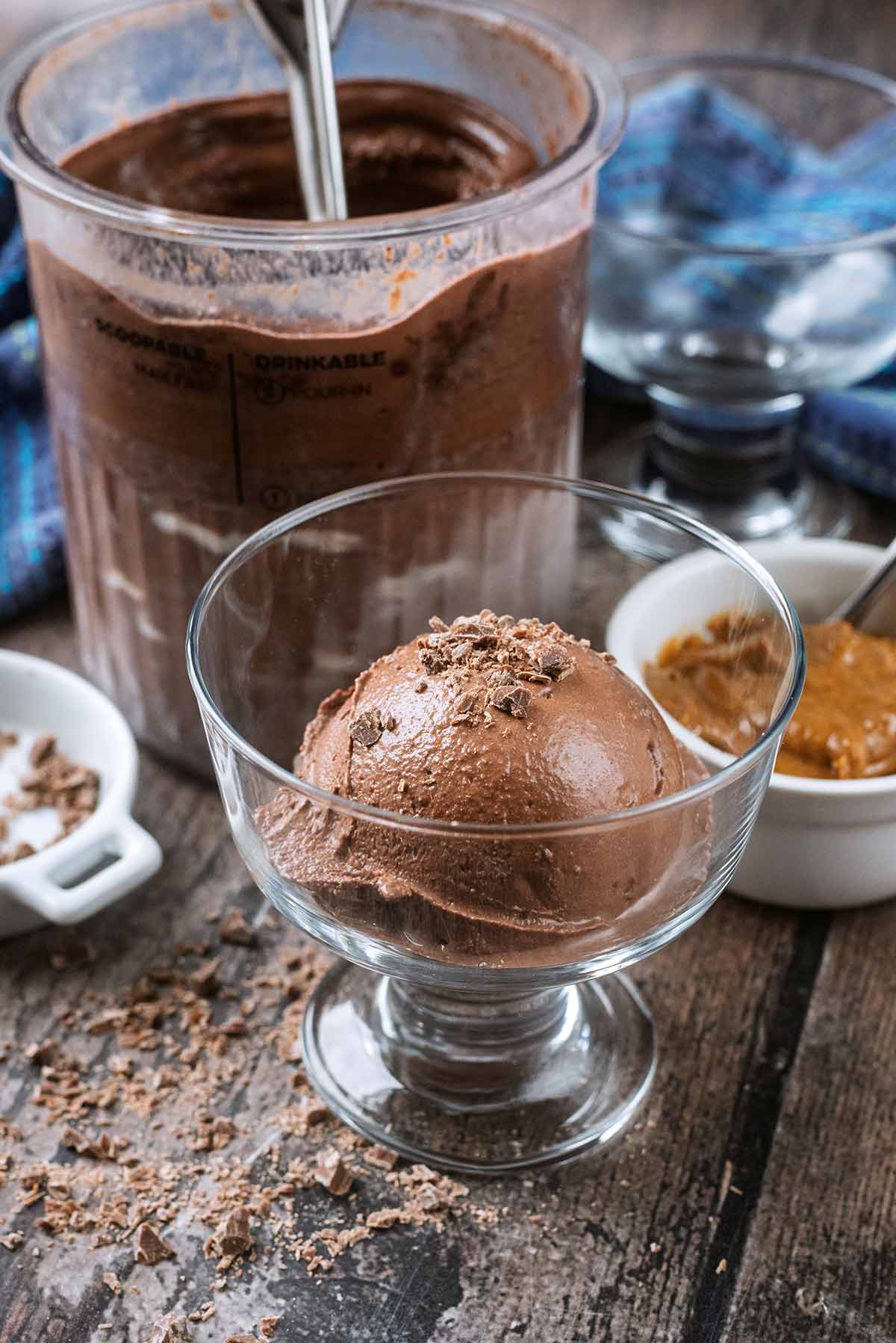 A ball of chocolate ice cream in a serving glass in front of a tub of more ice cream.