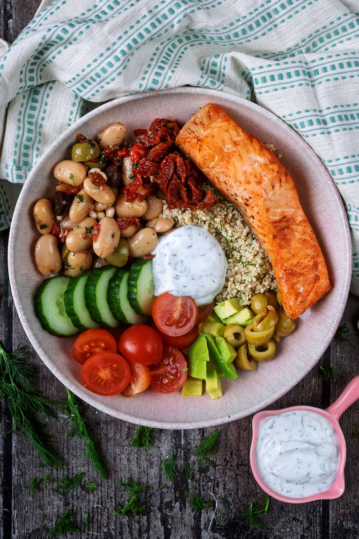 A bowl of salad vegetables, a cooked salmon fillet, cooked quinoa and a creamy dressing.