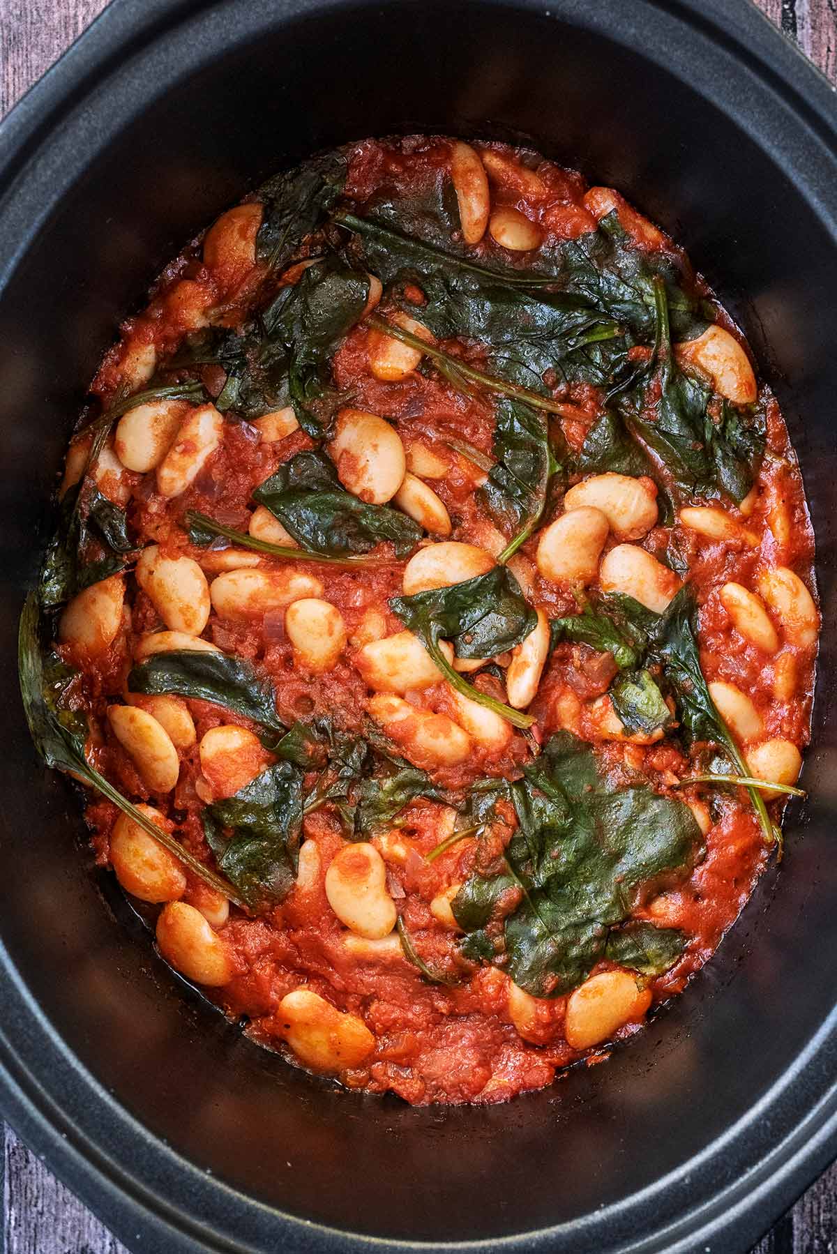 A slow cooker bowl containing butter beans and spinach in a tomato sauce.