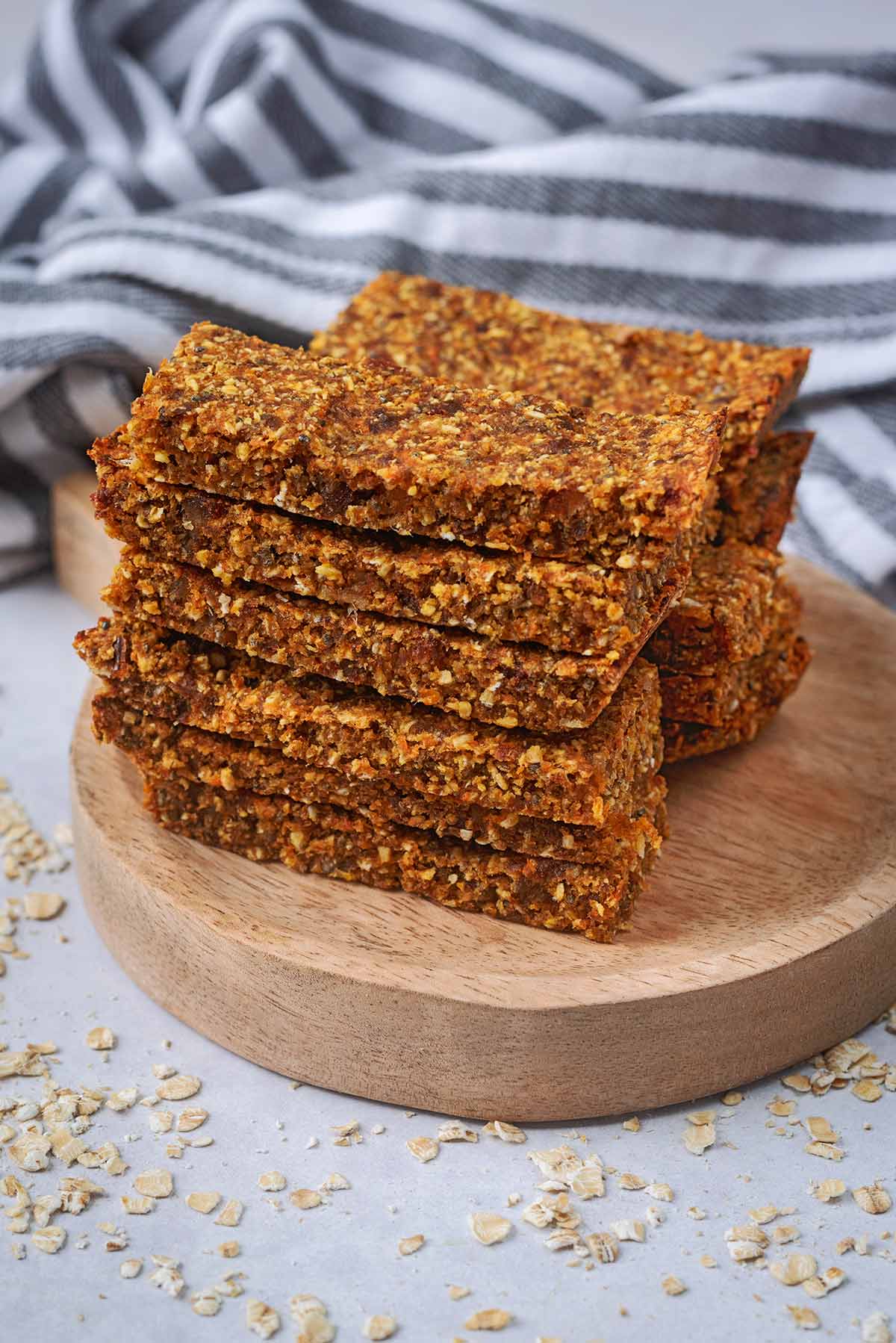 Oat bars piled up on a small wooden board with a striped towel in the background.