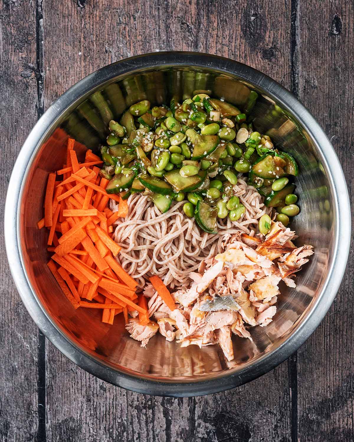 A mixing bowl containing flaked salmon, cooked noodles, carrot battens, edamame and diced cucumber.