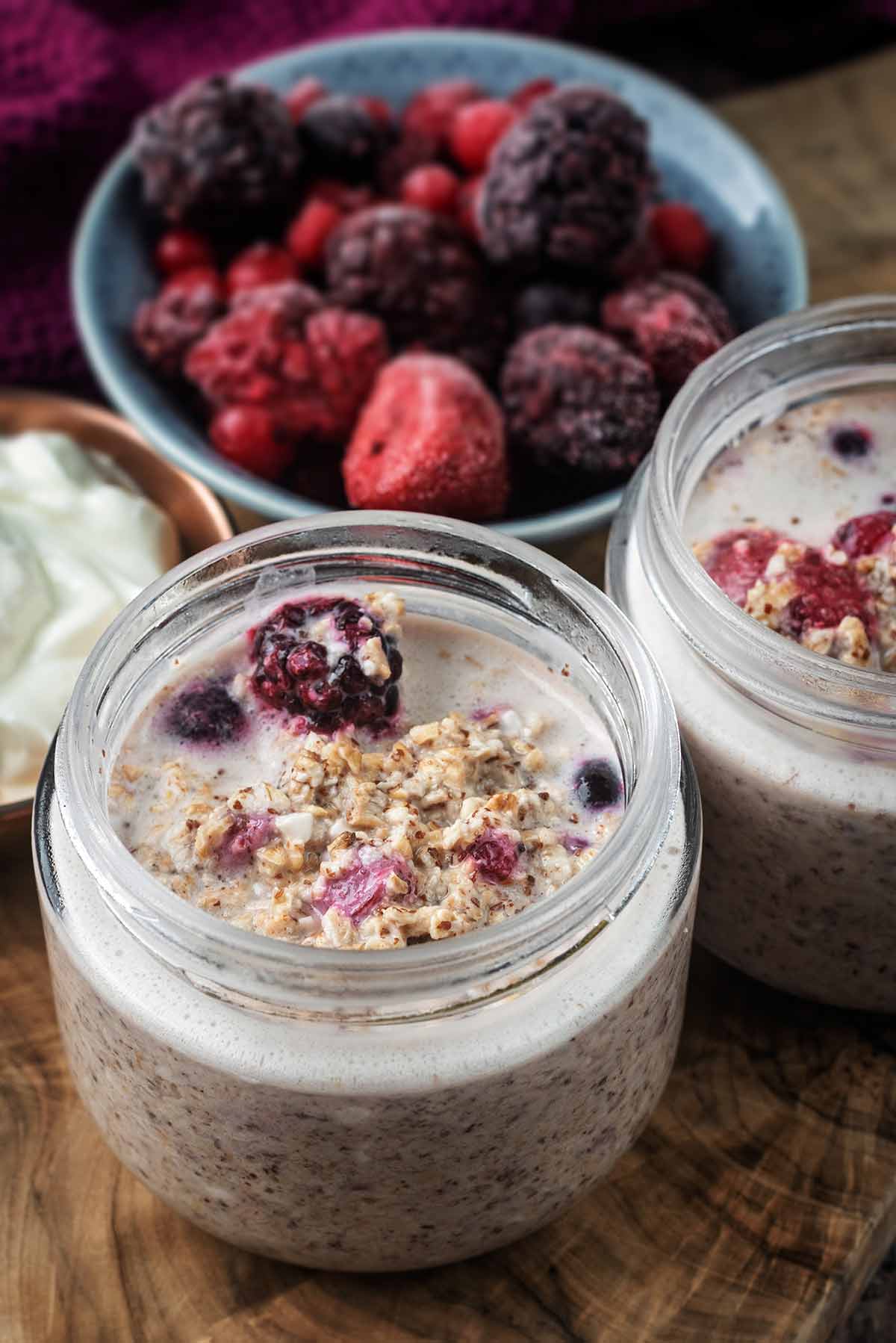Two jars of overnight oats next to a bowl of frozen berries.