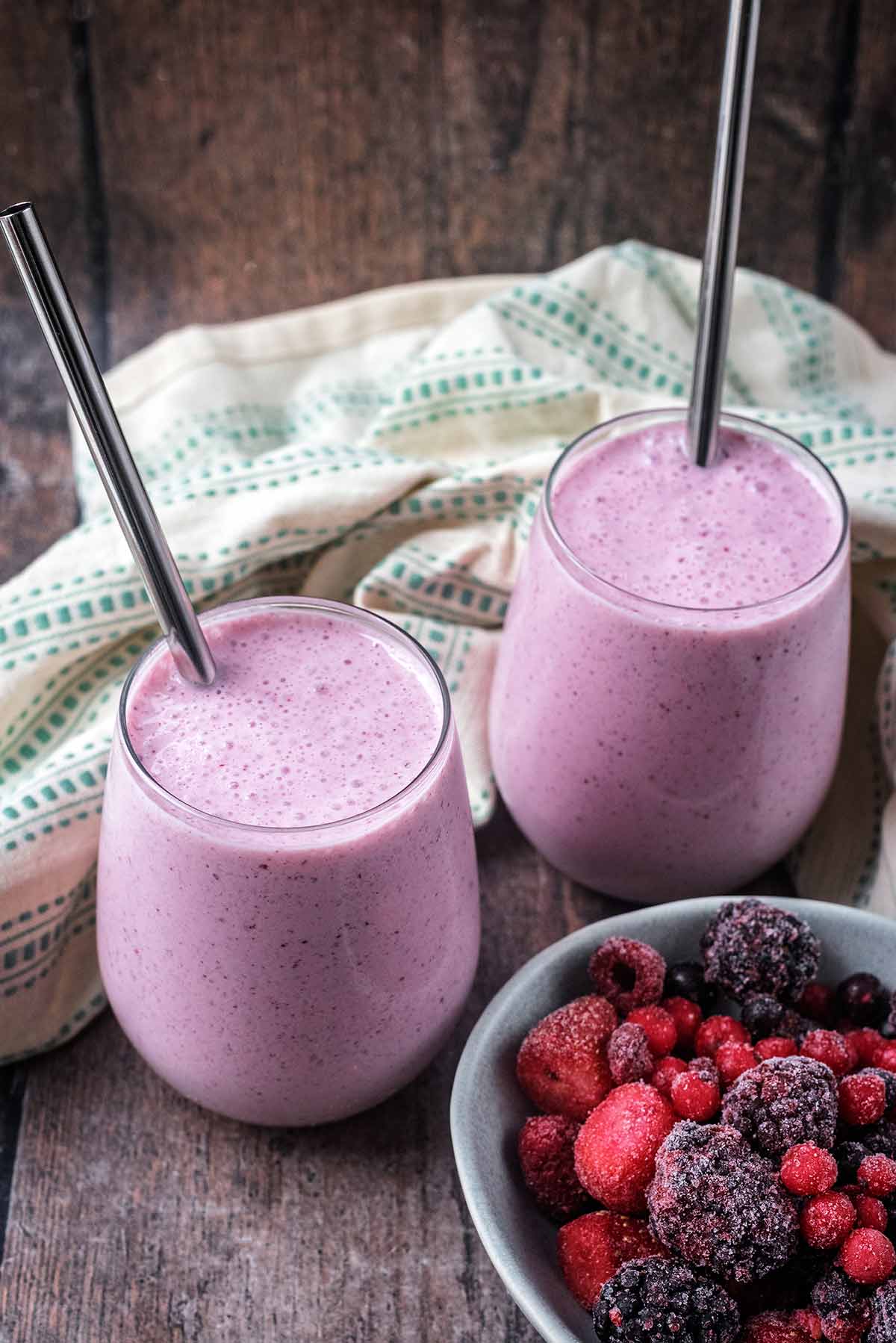 Two glasses of smoothie next to a towel and a bowl of berries.