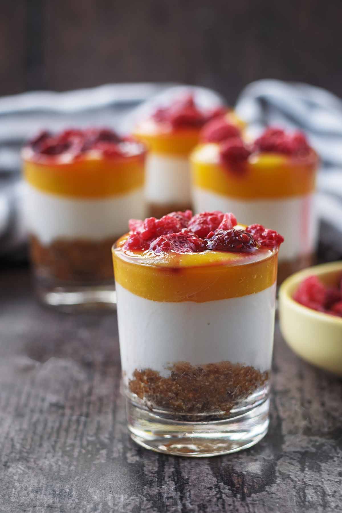 A small glass containing mango cheesecake with three more cheesecakes in the background.
