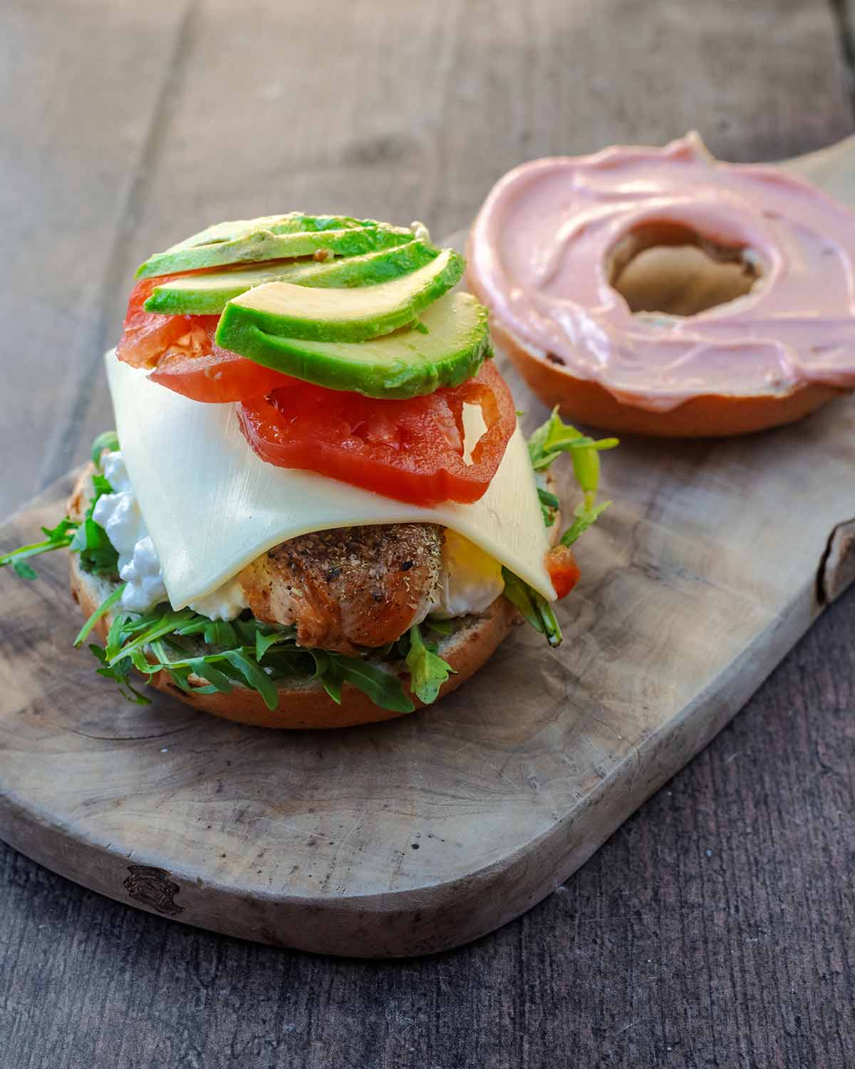 A bagel half topped with cottage cheese, lettuce, chicken, cheese slice, tomato and avocado. The other bagel half lies behind with pink sauce spread on it.
