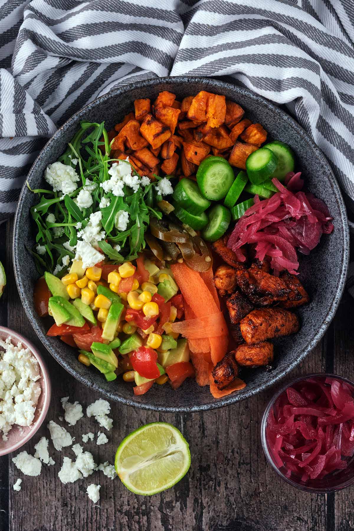 A bowl of cooked diced chicken, salad items and cheese, next to a lime half and some crumbled feta.